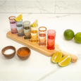 Set of shot glasses with colorful drinks on a wooden board, accompanied by limes and seasonings.